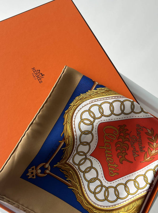 A partially unfolded Hermès - Silk Scarf by EXCHANGE, made of 100% silk with a red, gold, and blue pattern, rests atop the iconic orange box featuring the brand’s logo.