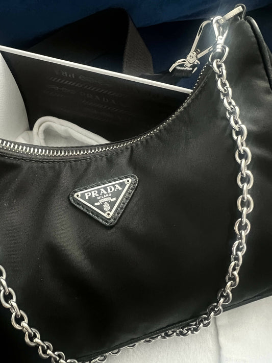 A close-up of the EXCHANGE Prada Re-Edition 2005 Re-Nylon Bag with a silver chain strap and front logo. Its practical design and saffiano leather trim are showcased by a book peeking out from the open bag.